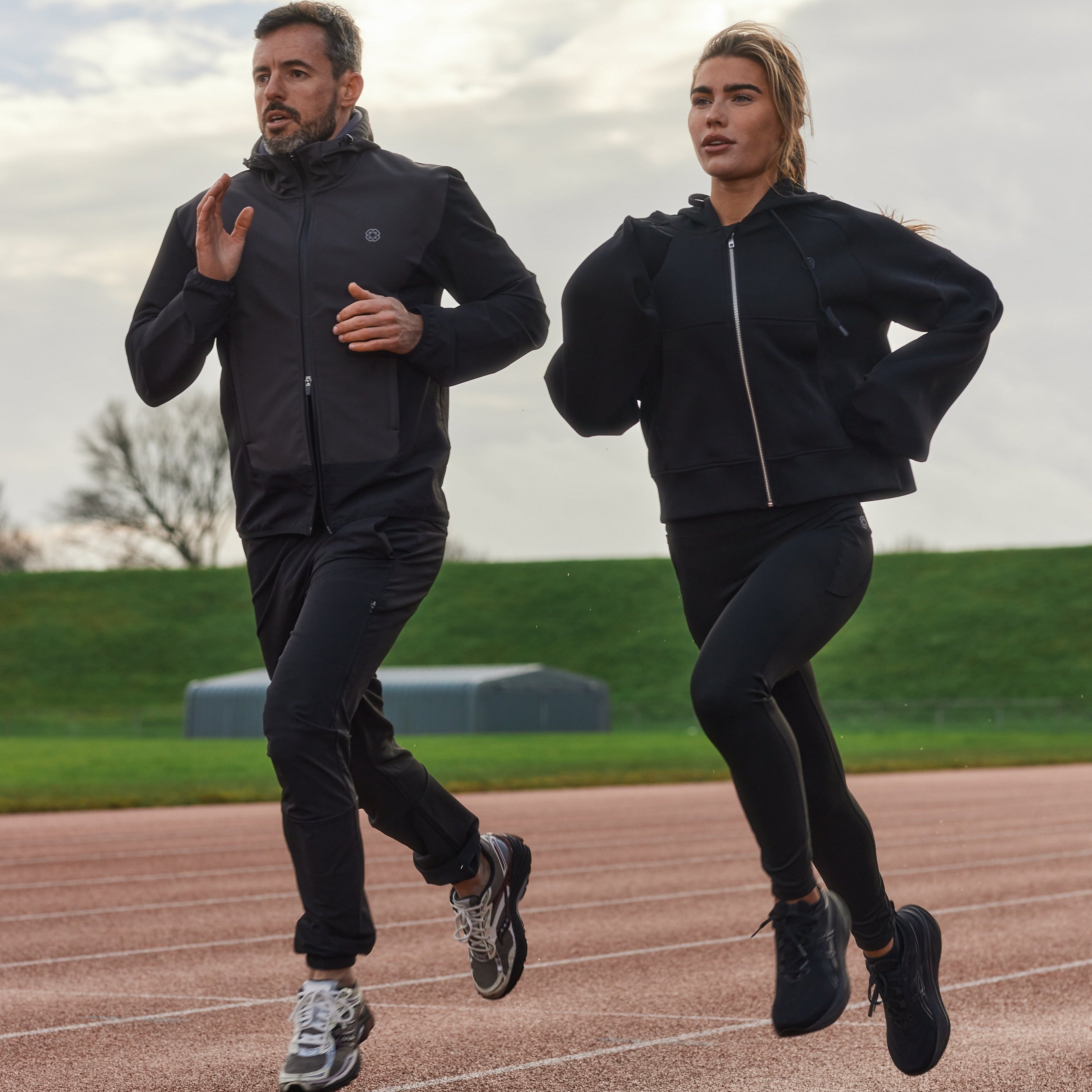 Two people running on a track wearing black athletic jackets and pants.